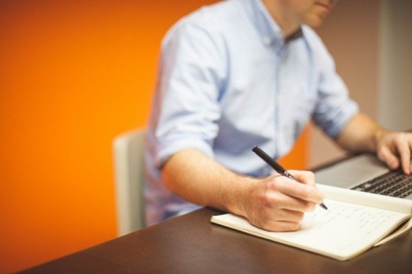 young-man-sitting-at-desk-using-laptop-and-writing-in-notebook